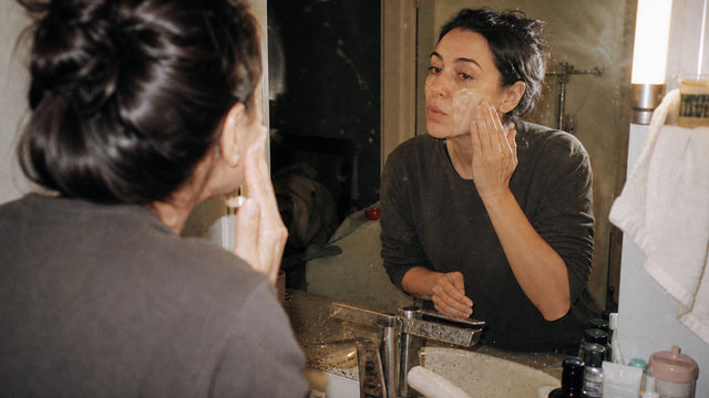 Adult using cleansing oil at a lived-in bathroom mirror in warm evening light